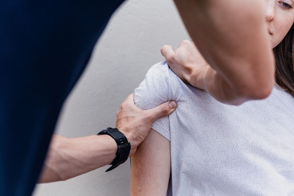 A person massaging the shoulder of another person wearing a white patterned shirt
