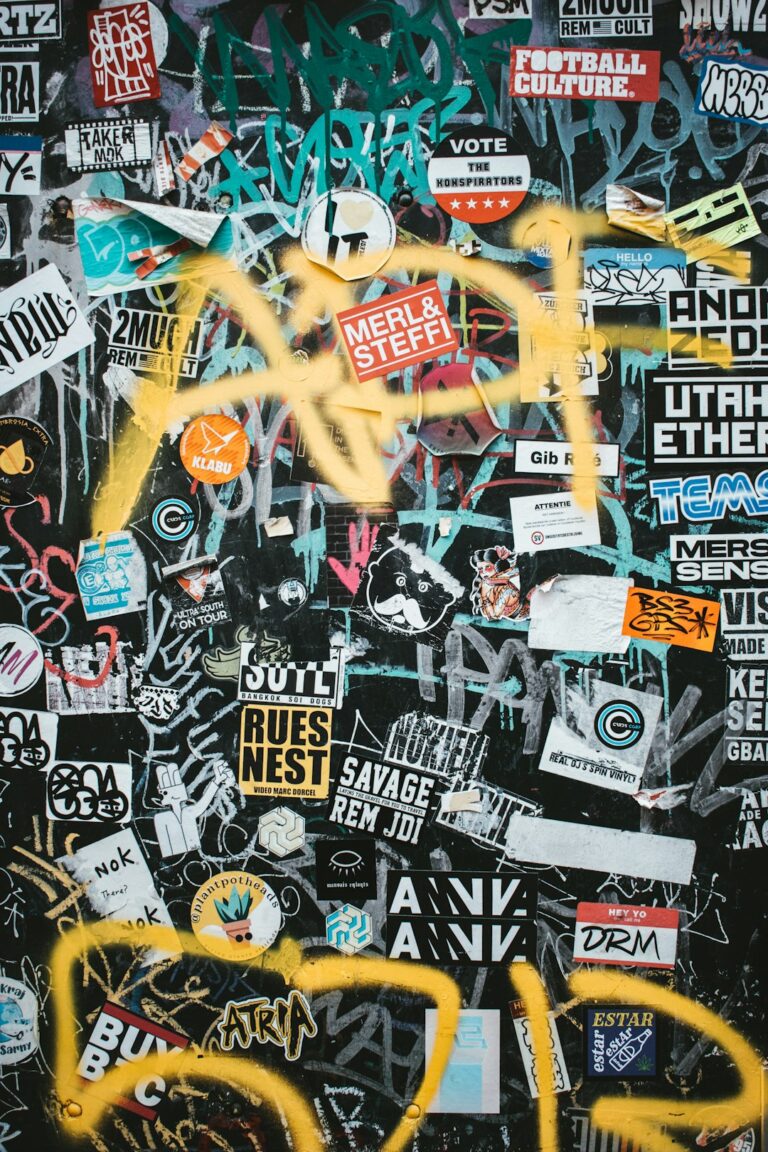 A dark wall covered in numerous colorful stickers and yellow spray paint graffiti